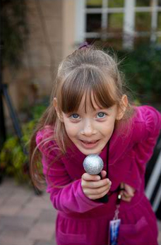 Young girl in a pink jacket holding a silver microphone, smiling at the camera outdoors.