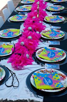 A festive outdoor table setting with pink feather boas, colorful plates, and scattered party favors along a long table.