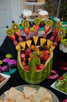 A watermelon carved into a decorative bowl filled with various fruits and garnished with fruit skewers, displayed on a party table.