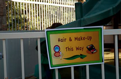 Directional sign for "hair & make-up" with an arrow, scissors, and a comb illustration, placed on a white fence outdoors.