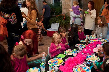 Children and adults at an outdoor birthday party with a table set for a meal and colorful decorations.
