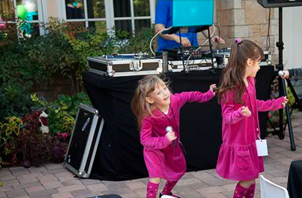 Two young girls in pink outfits dancing joyfully in front of a dj booth at an outdoor event.