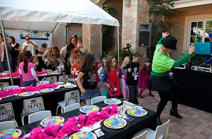 An outdoor children's party with kids dancing and adults watching. a table in the foreground displays colorful prizes and toys.