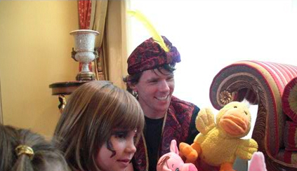 Two people smiling, one holding a stuffed duck, in a room with ornate decor. one person wears a colorful turban and a red patterned jacket.