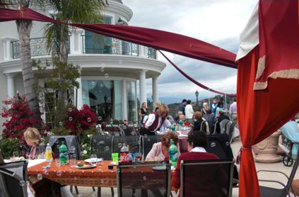 Outdoor dining event at a white villa with people seated at tables, a waiter serving, and an ocean view in the background.