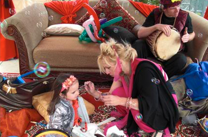 A woman paints a child's face at a festive event, while a man in a hat plays a drum on a decorated couch surrounded by colorful props.