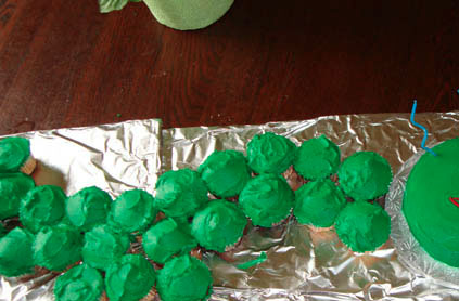 Green frosted cupcakes arranged in a line on a foil-covered table, with a green object on the left and a plate on the right.