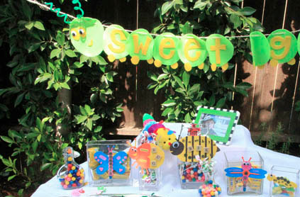 Colorful candy-themed party table setup outdoors with decorative banner, assorted candies in clear jars, and themed crafts on a white tablecloth.