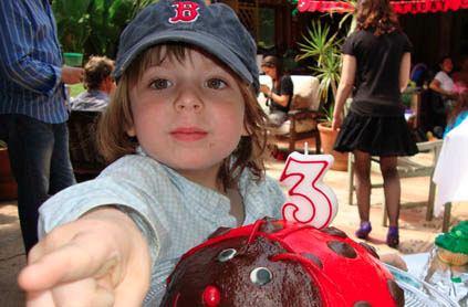 Young child in a baseball cap reaching towards the camera at a birthday party, with a cake decorated as a ladybug and a '3' candle in front.