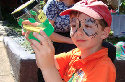 A boy with face paint resembling a spider web, wearing a red cap and orange shirt, holds a decorated jar at an outdoor event.