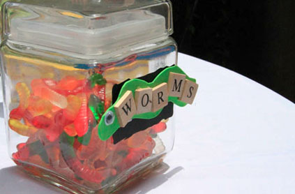 A clear square jar filled with colorful gummy worms, labeled with scrabble tiles spelling "worms" on its side.