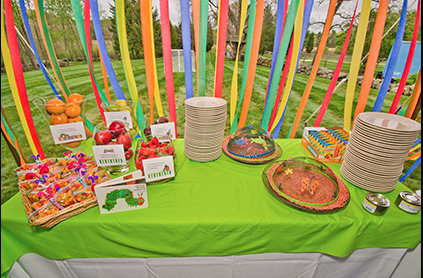 Colorful outdoor buffet table with snacks, plates, and ribbons hanging in a garden setting.