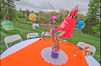 A colorful outdoor party table decorated with makeshift flower adornments, bright butterfly cutouts, and a large pink kite, set on a green lawn.