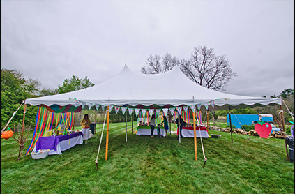 A large white tent with colorful ribbons at an outdoor event, with tables under the tent and a cloudy sky above.