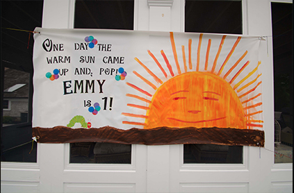 Banner hanging on a house featuring a painted smiling sun and text "one day the warm sun came up and pop! emmy is 1!" celebrating a first birthday.