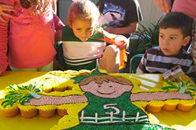 Children gathered around a table with a large birthday cake shaped like a cartoon girl in a green dress.