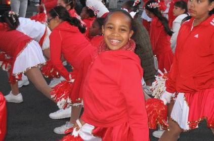 A young girl in a red hoodie and cheerleading skirt smiling at the camera during a parade, with other cheerleaders in the background.