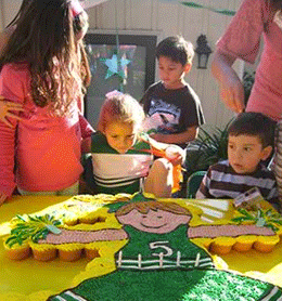 Children gather around a table with cupcake cake during a birthday party, the cake decorated with a name "lila.