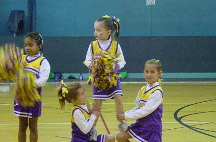 Young cheerleaders in purple and yellow uniforms performing a routine in a gymnasium, with one girl being lifted by her teammates.