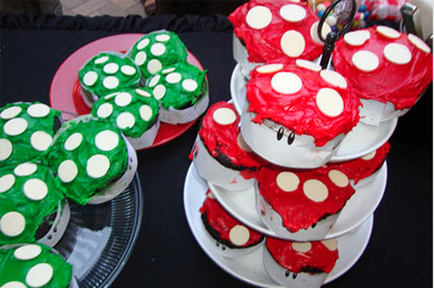 Stacked plates holding red and green frosted cupcakes decorated to resemble mushrooms, set on a table at an outdoor event.