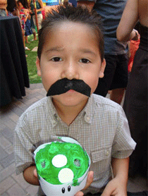Young boy with a fake mustache holding a green dessert in a cup, smiling at a party.
