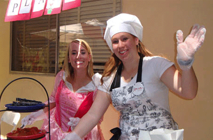 Two women in costumes, one dressed as a chef and the other in a pink dress, waving and smiling at an outdoor event.