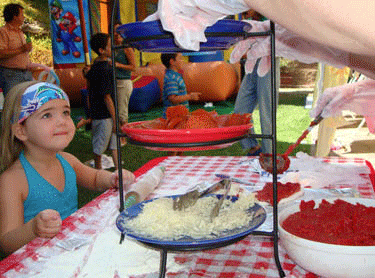 A young girl watches as a server prepares a plate of food at an outdoor party, with other children playing in the background.