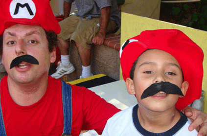 Two people wearing red hats and black mustaches resembling mario characters, one adult and one child, smiling at a party.