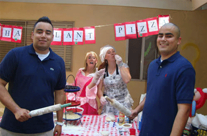 Three people smiling at an outdoor pizza-making party, with two holding rolling pins and one woman tossing dough under a "happy birthday pizza" banner.