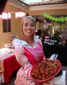 A woman wearing an apron smiles while holding a bowl of fried food at an outdoor party with decorations visible in the background.