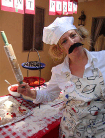 Woman dressed as a chef with a fake mustache, holding a paintbrush over a pizza on a table at a party, apron and chef hat on.