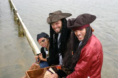 Three people dressed as pirates smiling by a wooden dock over a body of water, one holding a small wooden chest.