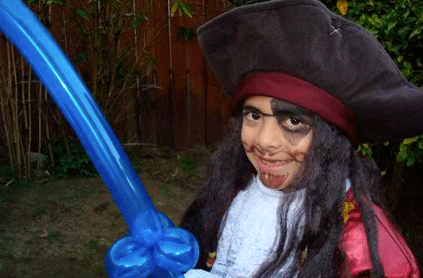 A child dressed as a pirate with a large hat and face paint, holding a blue balloon sword, standing in a backyard.