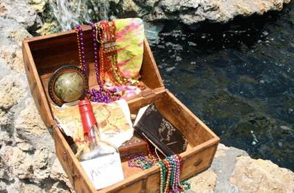An open treasure chest filled with colorful beads, a rolled map, a compass, and a bottle, set beside a rocky waterside.