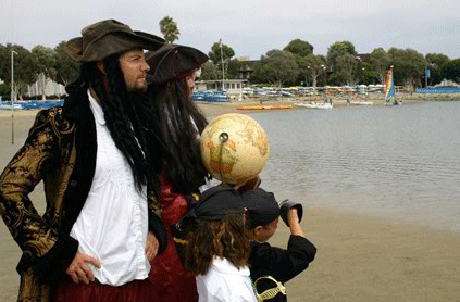 Two adults and a child in pirate costumes examine a globe by a lakeside with boats in the background.