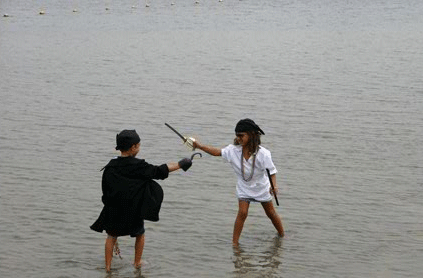 Two children play-fighting with sticks in a shallow river, one dressed as a pirate and the other in a black cloak.