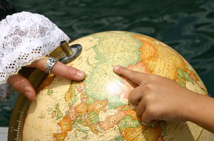 A person's hand pointing at a location on a globe, with another person holding a magnifying glass over it.