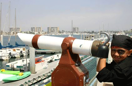 Man in a bandana smiling at the camera while looking through a harbor telescope, with boats and city skyline in the background.