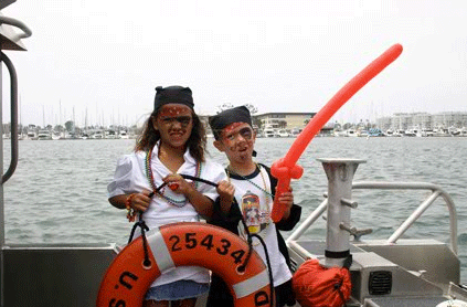 Two children dressed as pirates on a boat, holding a red balloon sword and standing next to an orange lifebuoy, with a marina in the background.