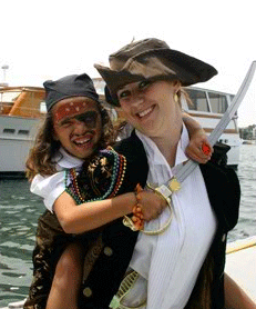 Two people dressed as pirates, smiling on a boat; one child and one adult, holding a plastic sword.