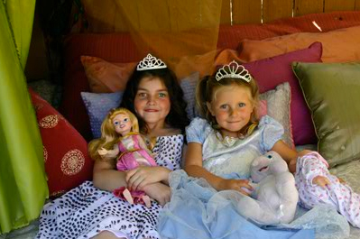Two little girls in princess dresses sit on a bed.