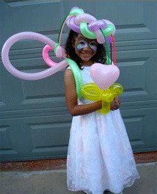 A young girl in a white dress smiling, wearing glasses and adorned with colorful balloon art including a large heart and a headpiece.