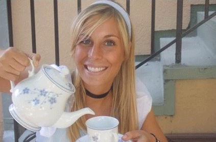 A smiling woman pours tea from a white floral teapot into a cup on a sunny balcony.