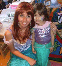 A woman and a young girl, both dressed in mermaid costumes, sit together in a colorful room filled with children's toys.