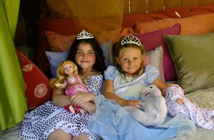 Two young girls wearing tiaras and holding dolls, sitting on a couch with colorful pillows.