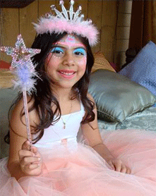 A young girl dressed as a fairy princess with a pink tutu, tiara, and holding a wand, sitting indoors with a joyful smile.