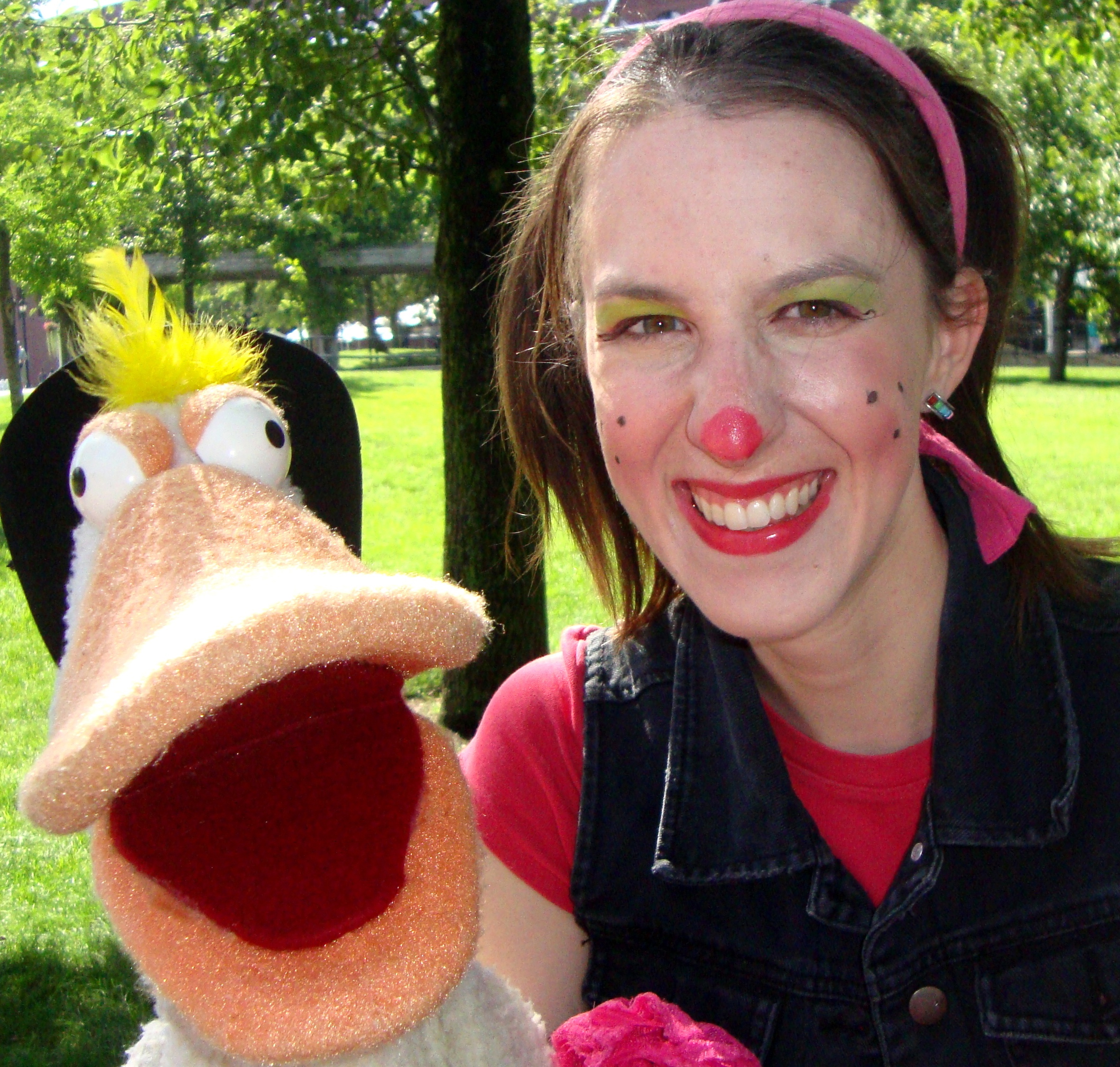 A woman with face paint smiles next to a duck puppet.
